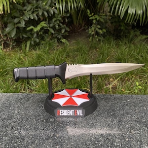 Op de afbeelding: Een zilverkleurig mes met een zwart handvat, gepresenteerd op een zwarte standaard met een rood-wit paraplu-logo en de tekst "RESIDENT EVIL". Het mes heeft een gekartelde rand en een gedetailleerd embleem.