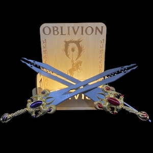 Könnte beinhalten: Zwei gekreuzte Fantasy-Schwerter mit verzierten goldenen Griffen und Schmuckakzenten, ausgestellt vor einem Holzschild mit dem Wort "OBLIVION" und einem Logo. Die Schwerter haben eine hellblaue Klinge.