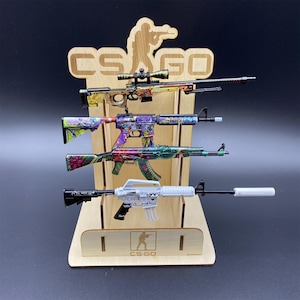 Op de afbeelding: Een houten displaystandaard met vier speelgoedgeweren met kleurrijke ontwerpen. De standaard heeft de tekst "CS:GO" en een silhouet van een schutter. De speelgoedgeweren hebben verschillende patronen.