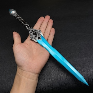 May include: A fantasy dagger with a bright blue blade and ornate silver hilt. The handle is wrapped in black material. The dagger is held in a hand against a dark background. The blade is approximately 10 inches long.