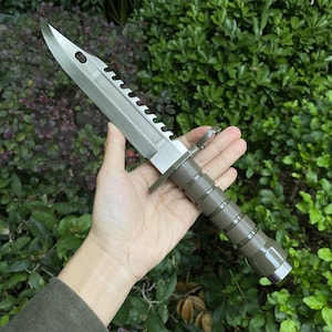 Puede incluir: Un cuchillo bayoneta plateado y verde oliva con borde dentado y mango texturizado. La hoja tiene un agujero cerca de la punta. El cuchillo se sostiene en una mano sobre un fondo de follaje verde.
