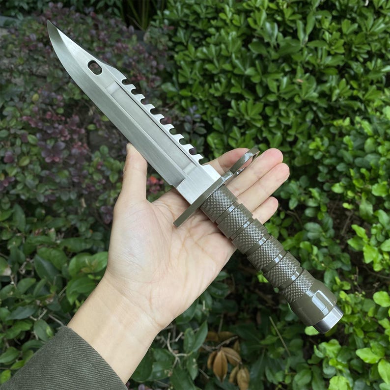 Puede incluir: Un cuchillo bayoneta plateado y verde oliva con borde dentado y mango texturizado. La hoja tiene un agujero cerca de la punta. El cuchillo se sostiene en una mano sobre un fondo de follaje verde.