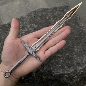 May include: A detailed miniature sword with a silver blade and a brown and cream-colored hilt. The hilt features intricate cross-shaped designs. The sword is held in a hand, showcasing its size and craftsmanship. A decorative item.