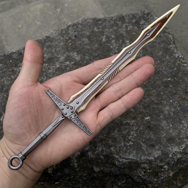 May include: A detailed miniature sword with a silver blade and a brown and cream-colored hilt. The hilt features intricate cross-shaped designs. The sword is held in a hand, showcasing its size and craftsmanship. A decorative item.