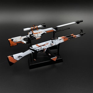 以下が含まれることがあります： 2つの玩具ライフル、スナイパーライフルとAK-47スタイルのライフルが表示されています。ライフルは白で、オレンジと黒のアクセントが付いています。スナイパーライフルにはスコープが付いています。どちらも暗い背景に対して黒いスタンドに取り付けられています。
