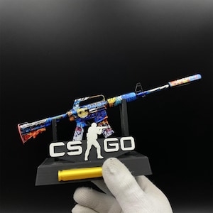 Pu&ograve; includere: Un fucile giocattolo colorato con un design blu, giallo e rosso, esposto su un supporto nero con la scritta "CS GO". Il fucile giocattolo &egrave; tenuto da una mano guantata.