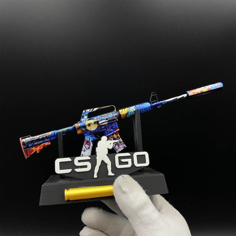 Pu&ograve; includere: Un fucile giocattolo colorato con un design blu, giallo e rosso, esposto su un supporto nero con la scritta "CS GO". Il fucile giocattolo &egrave; tenuto da una mano guantata.