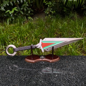 May include: A decorative metal kunai knife with a silver blade, a black handle, and a ring at the end. The blade features red and green accents. It is mounted on a wooden base.