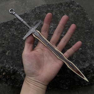 May include: A miniature sword with a silver-colored handle and crossguard, and a blade with a cream-colored center and brown edges. The sword has decorative details and a ring at the pommel.