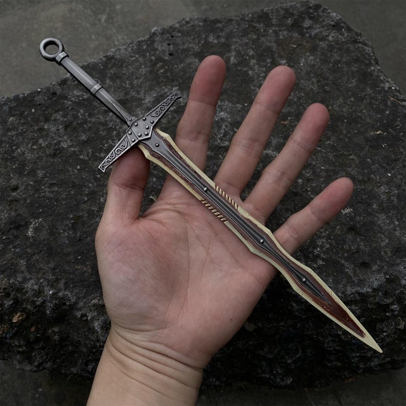May include: A miniature sword with a silver-colored handle and crossguard, and a blade with a cream-colored center and brown edges. The sword has decorative details and a ring at the pommel.