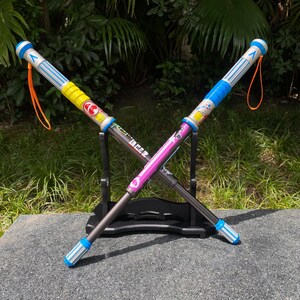 May include: Two colorful toy swords crossed on a black stand. One sword is pink and the other is yellow, with blue and silver accents. Each has an orange lanyard attached.
