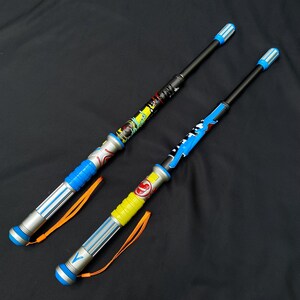 May include: Two toy wands with black shafts and colorful designs. One wand has a yellow and red handle, the other has a blue handle. Both wands have blue and silver accents and orange lanyards.
