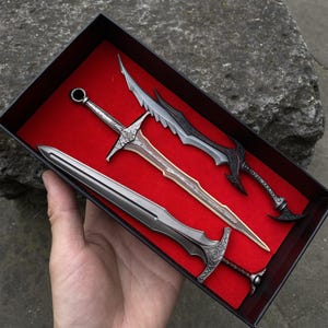 May include: A set of four miniature decorative swords in a black box with a red interior. The swords have silver, bronze, and black finishes. The swords are displayed in a box, with the blades facing up.