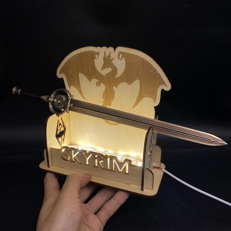 May include: A wooden Skyrim-themed display featuring a sword and a dragon silhouette. The base has the word "SKYRIM" cut out, with small lights illuminating the interior. The sword has a detailed hilt and a striped blade.