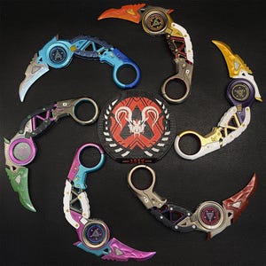 Könnte beinhalten: Eine Anordnung von sieben Karambit-Messern in verschiedenen Farben, darunter Blau, Orange, Lila und Grün, um ein kreisförmiges Emblem herum. Jedes Messer hat eine gebogene Klinge und einen Fingerring. Das Emblem ist schwarz, rot und weiß, mit dem Wort "APEX" unten.