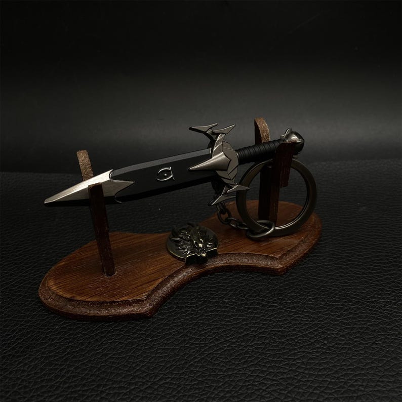 Puede incluir: Un llavero decorativo con forma de espada en miniatura, con una hoja negra y detalles plateados, exhibido en un soporte de madera. La espada presenta una empu&ntilde;adura detallada y est&aacute; unida a un llavero. Un peque&ntilde;o emblema ornamental descansa en el soporte.