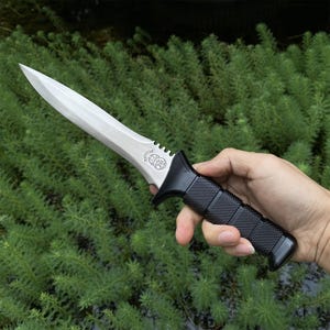 Puede incluir: Un cuchillo con una hoja plateada y un mango negro texturizado. El cuchillo tiene una hoja de doble filo y una guarda. El mango est&aacute; hecho de un material negro antideslizante. El cuchillo se sostiene sobre un fondo de follaje verde.