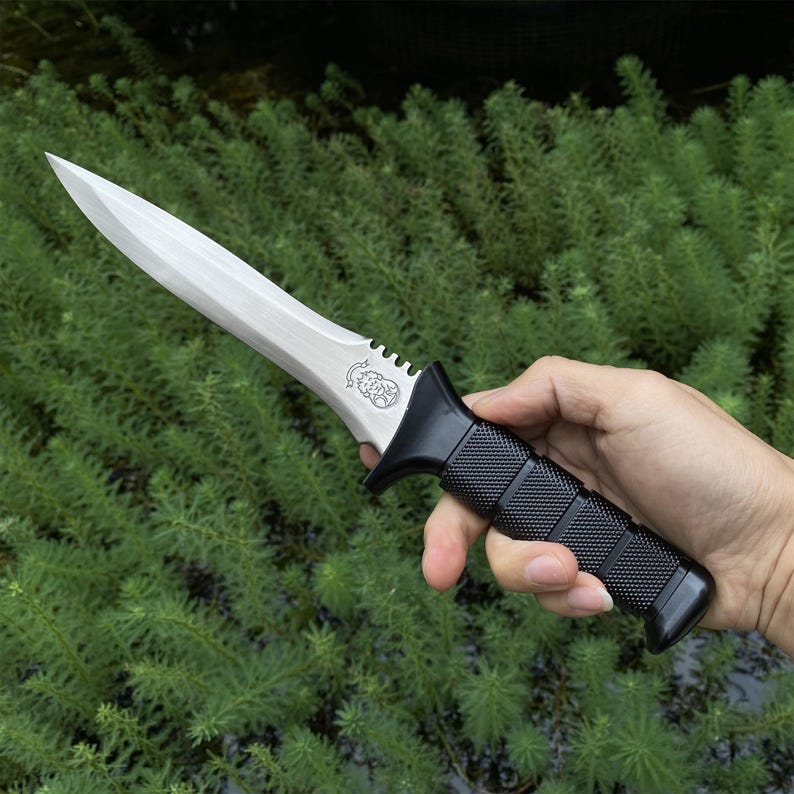 Puede incluir: Un cuchillo con una hoja plateada y un mango negro texturizado. El cuchillo tiene una hoja de doble filo y una guarda. El mango est&aacute; hecho de un material negro antideslizante. El cuchillo se sostiene sobre un fondo de follaje verde.