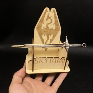Puede incluir: Una espada en miniatura con una hoja y empu&ntilde;adura plateadas, exhibida en un soporte de madera. El soporte presenta la palabra "SKYRIM" y un emblema de drag&oacute;n. El dise&ntilde;o general es de color marr&oacute;n claro.