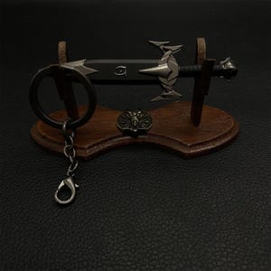 Puede incluir: Un llavero de espada en miniatura con un soporte de madera decorativo. La espada tiene una hoja negra y detalles plateados. Tambi&eacute;n se incluye un peque&ntilde;o emblema redondo y un llavero con cierre. El soporte es de madera marr&oacute;n oscuro.