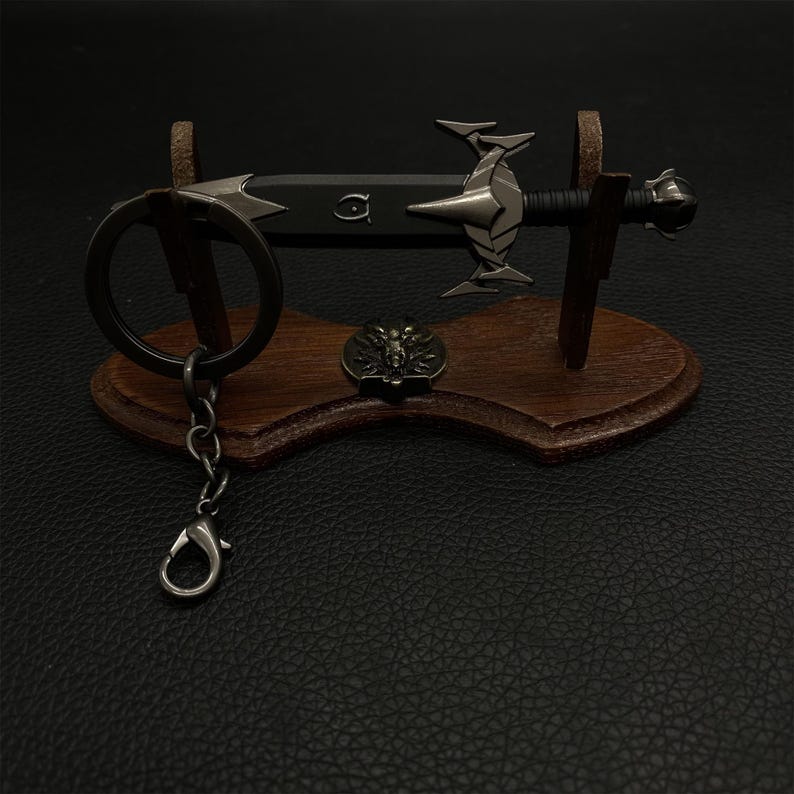 Puede incluir: Un llavero de espada en miniatura con un soporte de madera decorativo. La espada tiene una hoja negra y detalles plateados. Tambi&eacute;n se incluye un peque&ntilde;o emblema redondo y un llavero con cierre. El soporte es de madera marr&oacute;n oscuro.