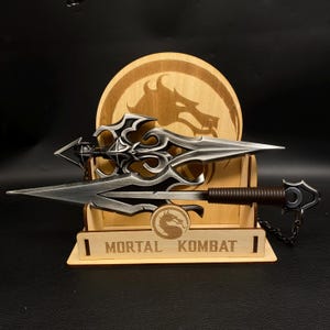 May include: A silver-colored spear with an ornate design, displayed on a wooden stand. The stand features a dragon logo and the text "MORTAL KOMBAT". The spear has a brown handle and a chain attached to the end.