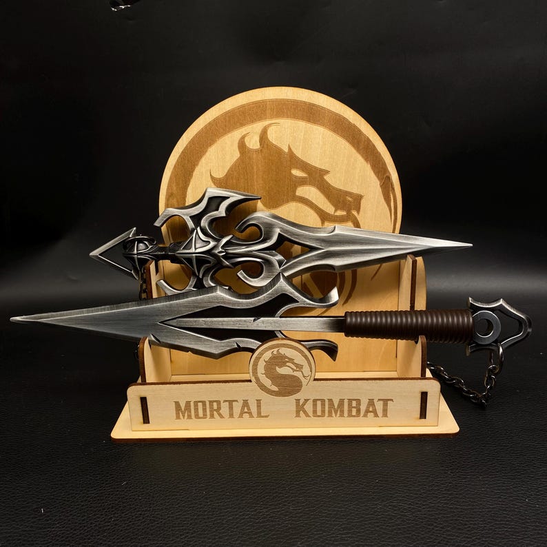May include: A silver-colored spear with an ornate design, displayed on a wooden stand. The stand features a dragon logo and the text "MORTAL KOMBAT". The spear has a brown handle and a chain attached to the end.