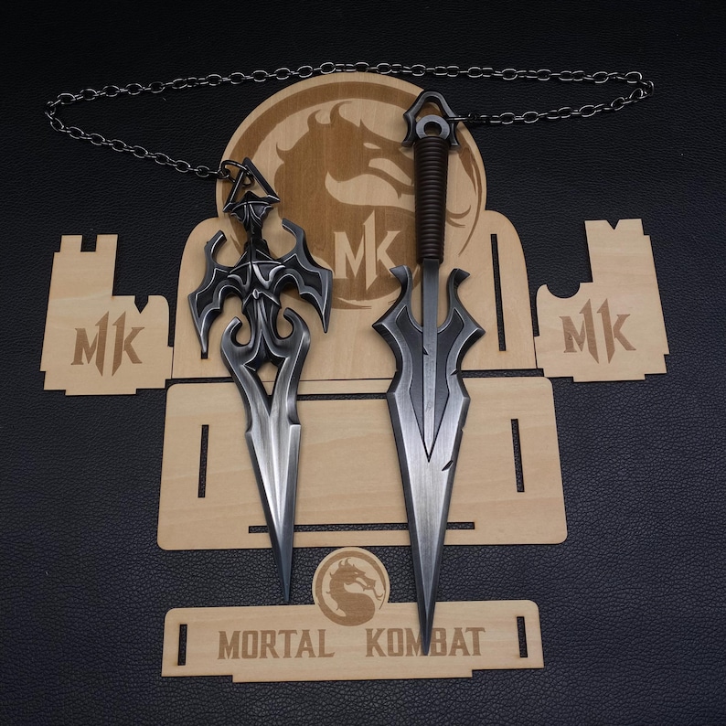 May include: Two metal dagger pendants with intricate designs, displayed on a wooden stand. One dagger has a dark handle and a chain, the other has a decorative hilt. The stand features the "Mortal Kombat" logo and "MK" initials.