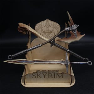 May include: A wooden display stand featuring crossed fantasy weapons from The Elder Scrolls V: Skyrim. The stand holds a bone club, a battle axe, and a sword. The stand is engraved with the game's title.