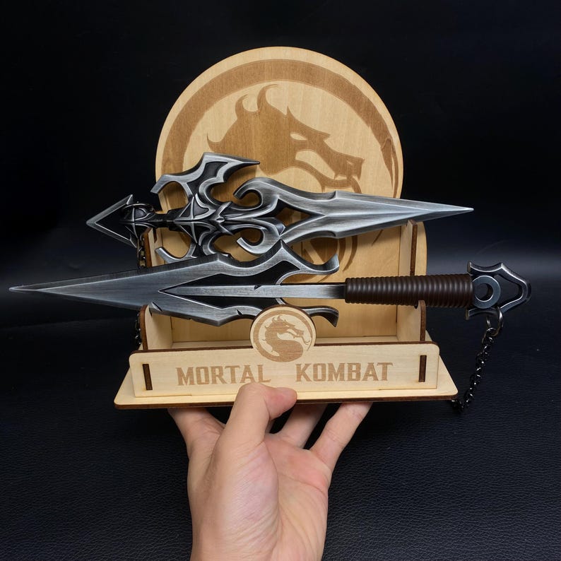 May include: A detailed display featuring two silver-colored, ornate blades with brown handles, presented on a wooden stand. The stand is engraved with the words "MORTAL KOMBAT" and includes a circular logo. A wooden backdrop with a dragon emblem completes the presentation.