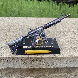 May include: A miniature rifle model featuring a dark metallic finish with a white claw mark pattern. The rifle is mounted on a black display stand labelled COUNTER-STRIKE with a yellow bullet-shaped object positioned in front of the base.