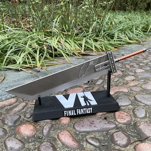 May include: A large, silver-colored Buster Sword replica from Final Fantasy VII, displayed on a black stand. The stand features the game's logo and the text "FINAL FANTASY". The sword has a brown handle and decorative accents.