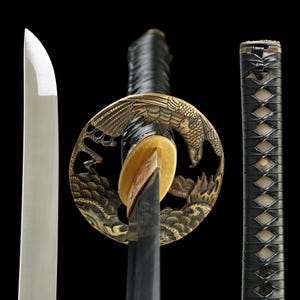 Peut inclure: Gros plan sur un katana japonais. La lame argentée contraste avec la garde noire et dorée ornée d'un motif d'aigle. Le manche est enveloppé d'un matériau noir.