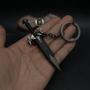 Puede incluir: Un llavero de espada en miniatura con una hoja negra y detalles plateados. El llavero presenta una empu&ntilde;adura y una guarda detalladas, unidas a una cadena y un gran llavero. El dise&ntilde;o recuerda a las armas de fantas&iacute;a.