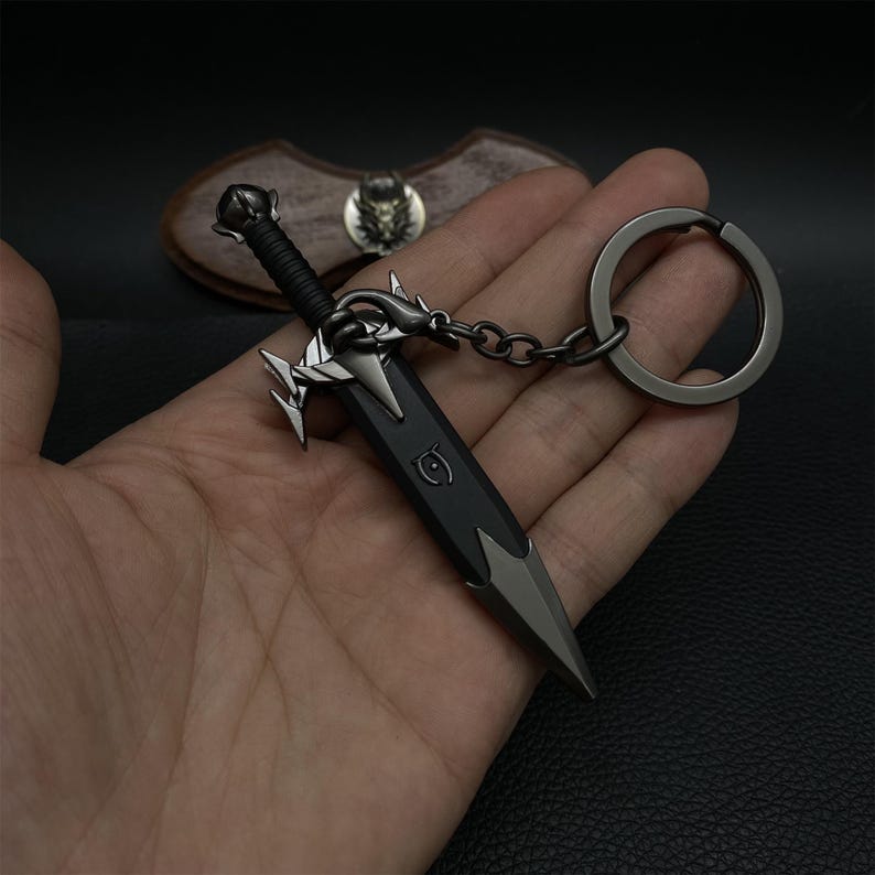 Puede incluir: Un llavero de espada en miniatura con una hoja negra y detalles plateados. El llavero presenta una empu&ntilde;adura y una guarda detalladas, unidas a una cadena y un gran llavero. El dise&ntilde;o recuerda a las armas de fantas&iacute;a.