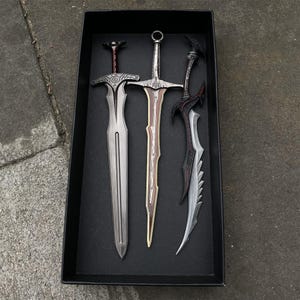 May include: Three decorative swords with ornate designs are displayed in a black box. The swords feature silver, brown, and black blades with detailed handles. The swords are arranged vertically, showcasing their unique shapes and styles.