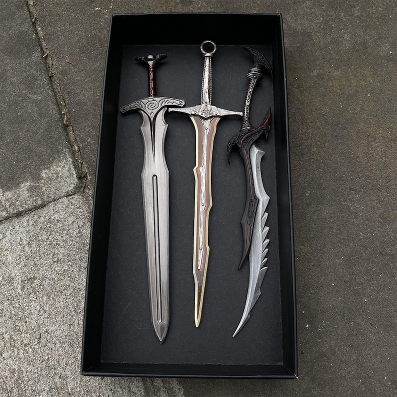 May include: Three decorative swords with ornate designs are displayed in a black box. The swords feature silver, brown, and black blades with detailed handles. The swords are arranged vertically, showcasing their unique shapes and styles.