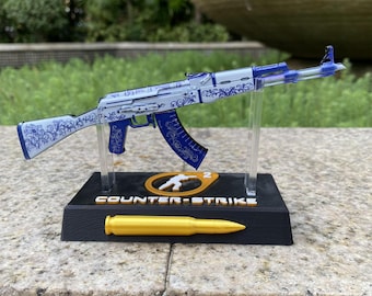 May include: A miniature rifle model featuring a white and blue floral pattern, mounted on a black display stand. The base displays the text COUNTER-STRIKE 2 with a gold-colored bullet replica positioned at the front of the stand.