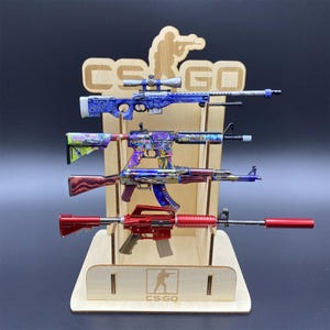 Op de afbeelding: Een houten displaystandaard met vier speelgoedgeweren in verschillende kleuren. De geweren zijn beschilderd in blauw, paars en rood. De standaard heeft de tekst "CS:GO" en een silhouet van een persoon met een geweer.