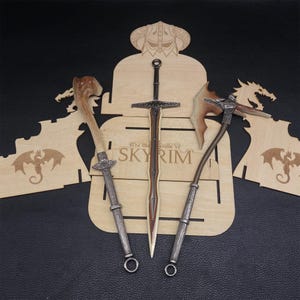 May include: A wooden display featuring three fantasy weapons: a sword, an axe, and a staff. The sword has a brown handle and silver crossguard. The axe has a brown handle and a silver head. The staff has a silver handle and a wooden top. The display has the text "The Elder Scrolls V SKYRIM".