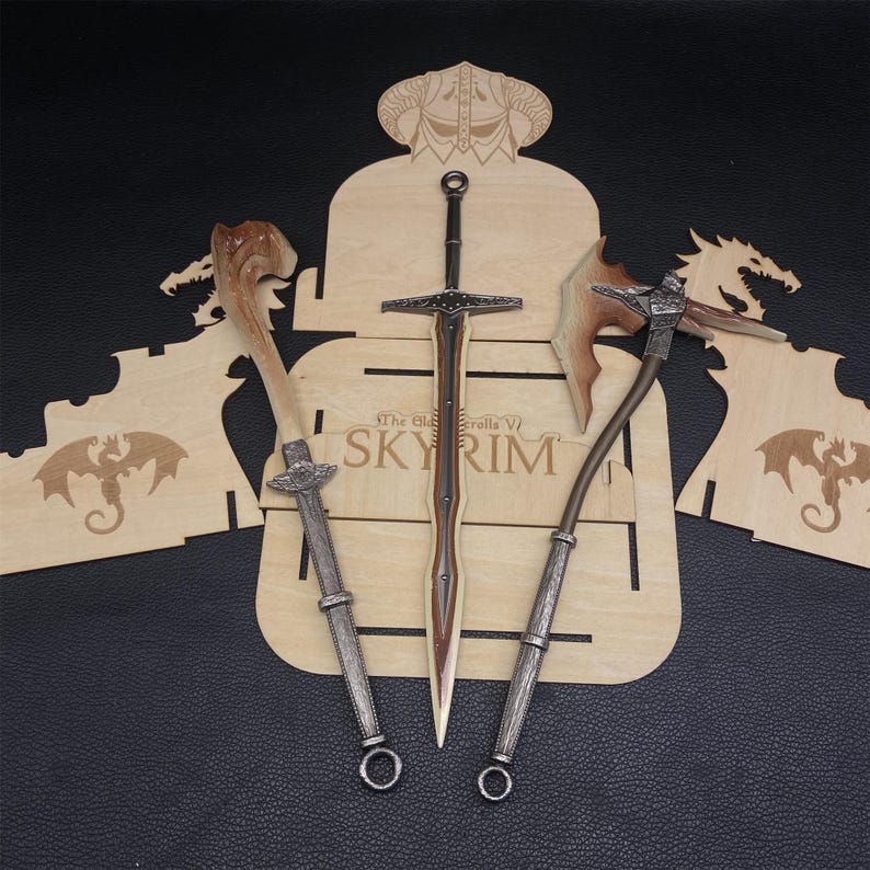 May include: A wooden display featuring three fantasy weapons: a sword, an axe, and a staff. The sword has a brown handle and silver crossguard. The axe has a brown handle and a silver head. The staff has a silver handle and a wooden top. The display has the text "The Elder Scrolls V SKYRIM".