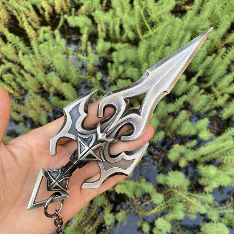 May include: A silver-colored dagger-shaped keychain with intricate, ornate designs. The blade is pointed, and the handle features a detailed pattern. The keychain is held in a hand against a backdrop of green foliage.