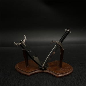 Puede incluir: Dos espadas en miniatura expuestas sobre una placa de madera. Una espada es negra con una empu&ntilde;adura plateada, y la otra tiene una hoja plateada y un mango negro. La placa es marr&oacute;n y tiene un borde decorativo.
