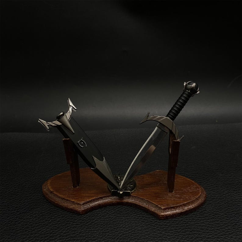 Puede incluir: Dos espadas en miniatura expuestas sobre una placa de madera. Una espada es negra con una empu&ntilde;adura plateada, y la otra tiene una hoja plateada y un mango negro. La placa es marr&oacute;n y tiene un borde decorativo.