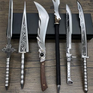 May include: An assortment of six decorative swords and knives with various blade shapes and handle designs. The blades are silver, and the handles feature silver, wood, and black wrapped designs. The collection is displayed on a wooden surface.