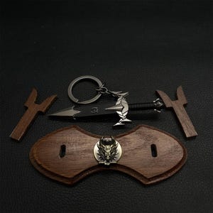 Puede incluir: Una colecci&oacute;n de art&iacute;culos de tem&aacute;tica fant&aacute;stica, que incluye un llavero de daga negra con un dise&ntilde;o de drag&oacute;n plateado, dos piezas de madera y una placa de madera con un emblema de lobo met&aacute;lico. Los art&iacute;culos est&aacute;n dispuestos sobre una superficie oscura.