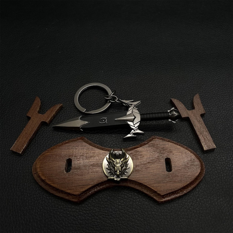 Puede incluir: Una colecci&oacute;n de art&iacute;culos de tem&aacute;tica fant&aacute;stica, que incluye un llavero de daga negra con un dise&ntilde;o de drag&oacute;n plateado, dos piezas de madera y una placa de madera con un emblema de lobo met&aacute;lico. Los art&iacute;culos est&aacute;n dispuestos sobre una superficie oscura.
