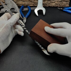 May include: A person is using a brown sanding block to smooth a metal object. The object is twisted and has a sharp point at one end. The person is wearing white gloves.