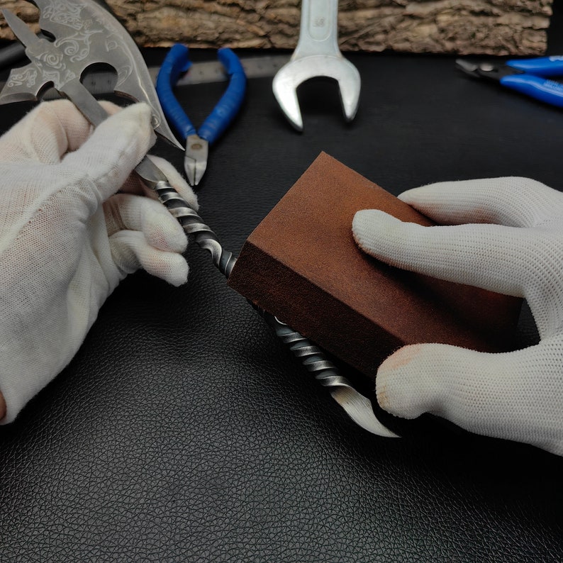 May include: A person is using a brown sanding block to smooth a metal object. The object is twisted and has a sharp point at one end. The person is wearing white gloves.