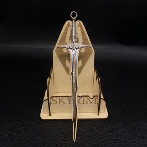 Puede incluir: Una espada detallada de color plateado con empu&ntilde;adura y hoja decorativas, expuesta sobre un soporte de madera. El soporte presenta la palabra "SKYRIM" y un logotipo. La espada tiene un anillo en la parte superior.